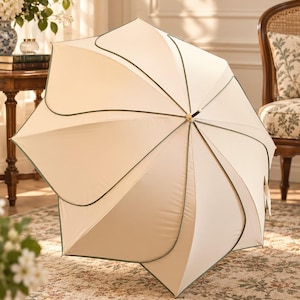 May include: A cream-colored umbrella with a scalloped edge design featuring dark green piping along the seams. The umbrella has a central metal shaft with a gold-toned tip and a curved handle, displaying a structured, multi-panel canopy shape.