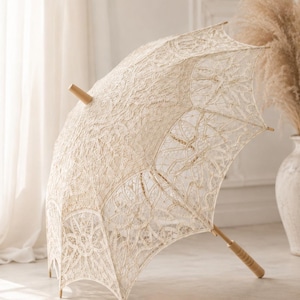 May include: A cream-colored lace parasol featuring an intricate floral pattern throughout the canopy. The parasol is constructed with a light-toned wooden handle and a matching wooden tip at the top, showcasing a delicate openwork design.