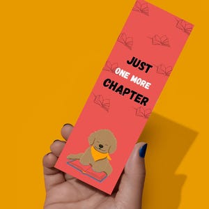 May include: A rectangular bookmark with a coral background featuring a repeating pattern of small black line-art books. The text JUST ONE MORE CHAPTER is printed in black and white. A cartoon brown puppy wearing a yellow bandana rests on a book.