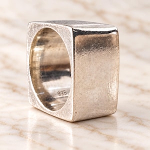 Op de afbeelding: Een dikke, vierkant gevormde zilveren ring met een getextureerde, matte afwerking. De binnenzijde van de band bevat een duidelijke gravure van het getal 925, wat de samenstelling van sterling zilver aangeeft van dit massieve, geometrische sieraad.