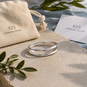 Puede incluir: Un anillo de plata de ley 925 pulida con un diseño de doble banda y una onda sutil. El anillo viene acompañado de una bolsa de tela beige con cordón y una pequeña tarjeta blanca, ambas impresas con el texto 925 STERLING SILVER.