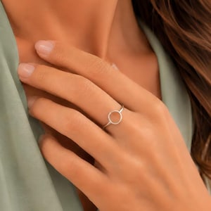 Puede incluir: Un anillo fino de plata pulida que presenta un pequeño diseño circular abierto en el centro. La banda es lisa y metálica, mostrando una forma de bucle simple y continua que se asienta en el dedo con un acabado brillante y reflectante.