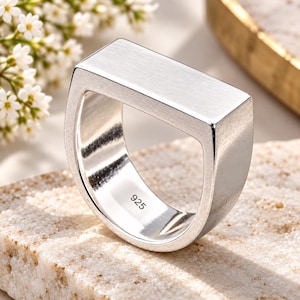 May include: A silver-toned ring featuring a flat, rectangular top surface and a smooth, brushed metal finish. The interior of the band is clearly stamped with the number 925, indicating the sterling silver composition of this jewelry piece.