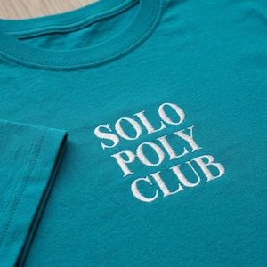 May include: A teal t-shirt featuring white embroidered text on the chest that reads SOLO POLY CLUB. The shirt has a crew neckline and short sleeves, with the embroidery centred on the front panel of the garment in a clean, sans-serif font.