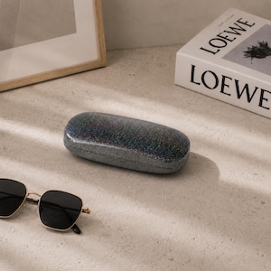May include: A dark, glittery, oval-shaped hard-shell glasses case with a textured finish. Nearby are black-lensed sunglasses with thin gold-toned metal frames. A white book with the word LOEWE printed in black text is visible in the background.