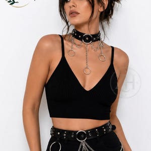 Puede incluir: Una gargantilla de cuero negro con anillos de metal plateado y cadenas colgantes, combinada con un cinturón de cuero negro a juego adornado con tachuelas plateadas, un anillo central y tres cadenas plateadas superpuestas en la cintura.