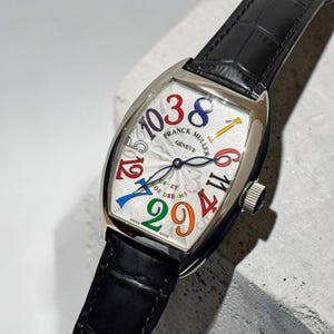 Op de afbeelding: Een Franck Muller Geneve polshorloge met een tonvormige zilveren kast en een zwarte lederen band. De witte wijzerplaat heeft grote, veelkleurige Arabische cijfers in een niet-opeenvolgende volgorde, met de tekst CRAZY OR DREAMS in het midden.