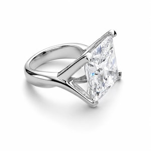 Op de afbeelding: Een zilverkleurige ring met een grote, vierkant geslepen heldere edelsteen in een zetting met vier pootjes. De band is glad en gepolijst en verbonden met de zetting via een V-vormige structuur die de steen boven de vinger verheft.