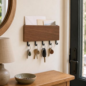 Può includere: Un organizer da parete rettangolare in legno con finitura marrone scuro. L'unità presenta una fessura superiore per contenere buste e cinque ganci in metallo nero lungo il bordo inferiore, che sostengono attualmente diversi mazzi di chiavi.