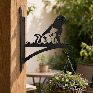 May include: A black metal wall-mounted plant bracket featuring a silhouette of a dog and a cat sitting together amidst flowers. The bracket has a curved hook at the end for hanging a basket and is attached to a wooden post with two screws.