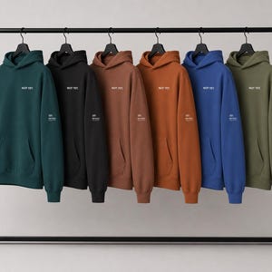 Puede incluir: Seis sudaderas con capucha y manga larga colgadas en un perchero de metal negro. Cada sudadera tiene un bolsillo tipo canguro, capucha y el texto "NOT YET" impreso en el pecho. Un pequeño gráfico con texto es visible en la manga izquierda.