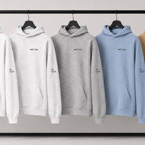 May include: Five hooded sweatshirts in white, light gray, heather gray, light blue, and tan hanging on a black rack. Each hoodie features the text NOT YET printed in black on the chest and a small matching text detail on the left sleeve cuff.