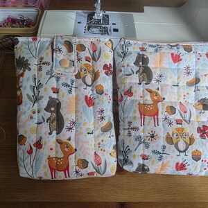May include: Two rectangular quilted fabric pouches featuring a repeating pattern of woodland animals including deer, owls, hedgehogs, and bears, interspersed with acorns and red flowers. One pouch has a flap with a snap closure, while the other is zippered.
