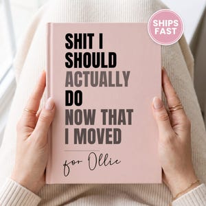 May include: A light pink hardcover notebook with black and dark gray text that reads: SHIT I SHOULD ACTUALLY DO NOW THAT I MOVED for Ollie. The notebook has a smooth, matte finish and a rectangular shape, held upright against a neutral background.