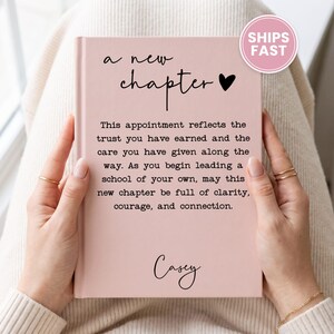 May include: A light pink hardcover journal featuring the black text 'a new chapter' with a heart icon, followed by a paragraph about leadership and the name 'Casey' in script font at the bottom of the front cover.