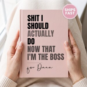 May include: A light pink hardcover notebook with black and dark gray text that reads: SHIT I SHOULD ACTUALLY DO NOW THAT I'M THE BOSS for Dana. The notebook features a clean, minimalist design with a matte finish and a circular pink badge in the corner.
