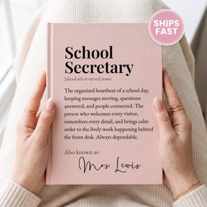 May include: A light pink hardcover book with black text that reads School Secretary, followed by a definition and the phrase Also known as: Mrs. Lewis. The cover features a dictionary-style entry describing the role of a school secretary.