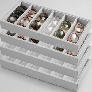 May include: A stack of four light gray rectangular storage trays with multiple interior compartments. The top tray contains various pairs of sunglasses with different frame colors and lens tints. The text LuxeUSA is visible on the tray.