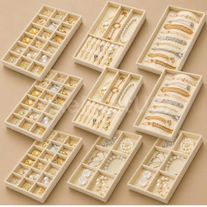 May include: A collection of nine beige velvet jewelry organizer trays arranged in a grid. The trays contain various gold-toned rings, earrings, necklaces, and bracelets, some featuring white pearls or clear crystals, organized into small compartments.