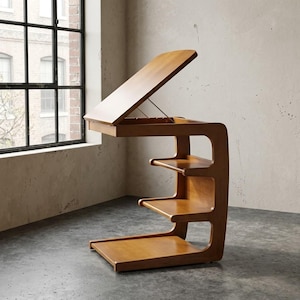 May include: A light brown wooden side table with a C-shaped frame, featuring an adjustable, tilting top surface supported by a metal hinge and two integrated lower shelves for storage, constructed with smooth, curved edges and a natural wood grain finish.