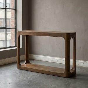 May include: A rectangular console table crafted from medium-brown wood. The piece features a smooth, flat top surface supported by two solid side panels with large, rounded rectangular cutouts, creating an open frame design with a base shelf.