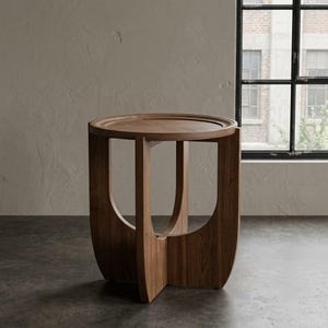 May include: A round side table crafted from medium-toned brown wood, featuring a circular tabletop with a raised edge and a base composed of three curved, vertical wooden panels that intersect to provide structural support for the surface.