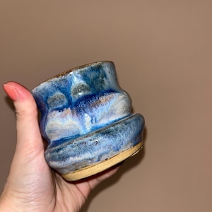 May include: A ceramic cup with a wavy, rippled silhouette and a glossy glaze featuring blended shades of deep navy, royal blue, and light sky blue. The base of the cup is made of unglazed, textured tan clay, contrasting with the smooth, colourful upper body.