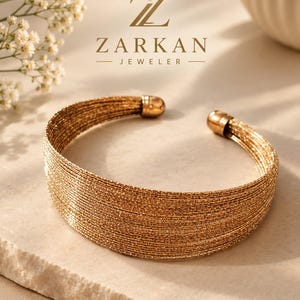 May include: A gold-toned cuff bracelet composed of multiple thin, textured wire strands arranged in a wide, parallel band. The bracelet features rounded, polished end caps and is positioned below the ZARKAN JEWELER logo with a diamond icon.