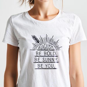 Könnte beinhalten: Ein weißes kurzärmeliges T-Shirt mit einem schwarzen Grafikdruck, der ein Sonnenstrahlen-Design mit einer Weizenähre zeigt. Der Text lautet: AUGUST. BE BOLD. BE SUNNY. BE YOU. Das Motiv ist mittig auf der Brust des T-Shirts mit Rundhalsausschnitt.