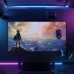 May include: A rectangular desk mat featuring a printed landscape illustration of a character standing on a rocky cliff overlooking a vast valley at sunset. The mat is positioned on a dark desk surface alongside a black gaming controller, a black computer mouse, and black over-ear headphones with blue light accents.