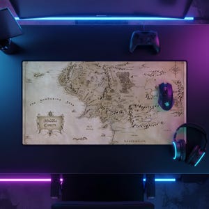 May include: A rectangular desk mat featuring a detailed, vintage-style map of Middle-earth with beige parchment-coloured background, black ink illustrations of mountains and forests, and the text Middle Earth prominently displayed in a decorative frame.