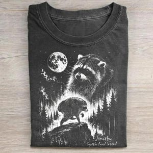 May include: A charcoal gray crew neck t-shirt featuring a white graphic print of a raccoon standing on a rock in a forest under a full moon. The text "Jimothy Seattle feral legend" is printed in white at the bottom right of the graphic design.