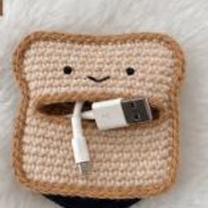May include: A square-shaped crochet pouch designed to resemble a slice of toasted bread with a light tan center and a darker brown crust border. The pouch features a simple embroidered face with two black eyes and a small smile, holding a white USB cable.