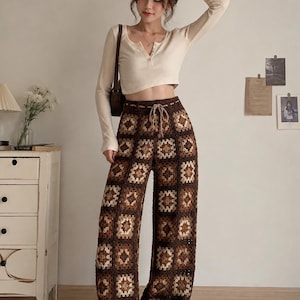 May include: A long-sleeved, cream-colored ribbed henley top paired with high-waisted, wide-leg crochet pants. The pants feature a repeating granny square pattern in shades of dark brown, medium brown, and beige, finished with a drawstring waist.