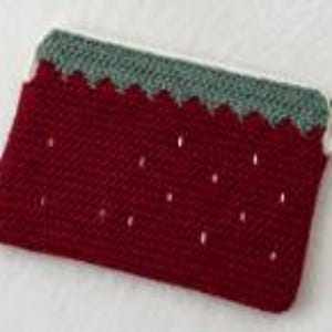 May include: A rectangular crochet pouch featuring a deep red body with small white embroidered accents resembling seeds. The top edge is finished with a scalloped green border and a white zipper closure, creating a strawberry-inspired design.