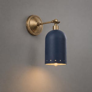 May include: A wall-mounted lamp featuring a matte navy blue dome-shaped shade with a row of small circular perforations along the bottom edge. The fixture includes an antique brass finish circular wall plate and an adjustable metal arm.