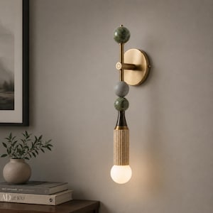 May include: A wall sconce featuring a vertical brass rod mounted to a circular brass base. The rod is adorned with two green marble spheres, one grey sphere, and a fluted cylindrical wooden section, terminating in a glowing white bulb.