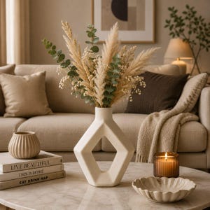 May include: A matte off-white ceramic vase with a geometric diamond-shaped cutout center, holding a dried floral arrangement of pampas grass, eucalyptus stems, and baby's breath, positioned on a marble surface with books and a candle.