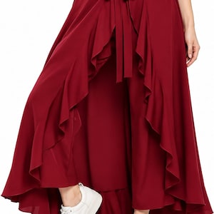 May include: A deep red wrap-style skirt with a high-low hemline, featuring cascading ruffles along the front opening and a matching fabric tie at the waist. The garment is paired with white sneakers, creating a layered look with a flowing silhouette.