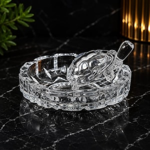 May include: A clear glass ashtray featuring a faceted, geometric rim design and a decorative clear glass turtle figurine positioned in the center. The turtle has a detailed shell pattern and is integrated into the circular base of the ashtray.