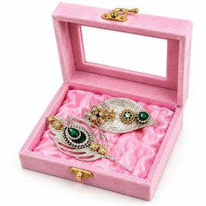 May include: A pink velvet jewelry box with a clear lid and gold latch contains two silver-toned brooches. One brooch is shaped like a peacock feather with a dark green teardrop stone, and the other is a leaf shape with green accents.