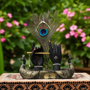 May include: A decorative sculpture featuring two black hands holding a gold flute, positioned behind a large peacock feather. The base is a gold-toned structure with two peacock figures on either side, adorned with small silver-coloured accents.