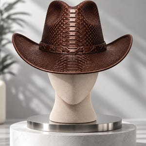 Può includere: Un cappello da cowboy marrone scuro caratterizzato da una trama a pelle di serpente sulla corona e sulla tesa. Il cappello include una fascia marrone abbinata con un dettaglio intrecciato attorno alla base della corona, esposto su un manichino.
