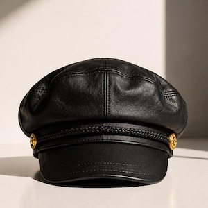 May include: A black leather baker boy cap featuring a structured crown with stitched paneling, a short curved brim, and a decorative braided cord trim across the front. The cap is accented with two small, circular gold-toned metal buttons on each side.