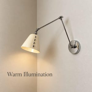 Può includere: Una lampada da parete con finitura in cromo lucido e braccio articolato regolabile. Il paralume conico è color crema e presenta una fila verticale di quattro piccoli rivetti argentati. La lampada emette luce, etichettata come Warm Illumination.