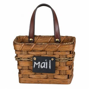 May include: A brown woven basket with a rectangular shape, featuring a dark brown leather handle attached with brass-colored rivets. A small black rectangular sign is attached to the front with the word "mail" written in white chalk-style lettering.