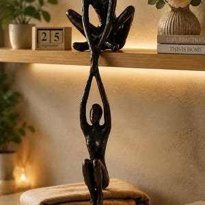 Pode incluir: Uma escultura em tom de bronze escuro com duas figuras humanas abstratas. Uma figura está sentada numa prateleira enquanto segura as mãos de uma segunda figura que pende verticalmente, com os joelhos dobrados em direção ao peito.