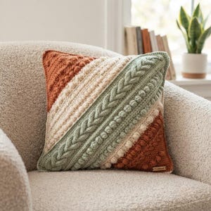 Puede incluir: Un cojín cuadrado con un patrón de rayas diagonales en hilo color naranja óxido, crema y verde salvia. El diseño incorpora puntos de ganchillo texturizados, incluyendo bodoques, trenzas y filas acanaladas, con una pequeña etiqueta de madera.