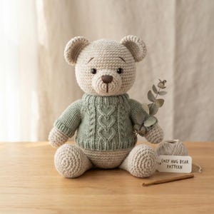 May include: A beige crocheted teddy bear wearing a sage green knitted sweater with a cable knit heart pattern. The bear holds a sprig of eucalyptus. Beside it sits a ball of yarn, a wooden crochet hook, and a tag labeled COZY HUG BEAR PATTERN.