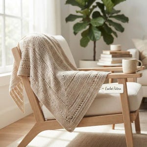 May include: A beige crochet blanket with a textured, openwork stitch pattern draped over a light-toned wooden chair. A small white rectangular tag attached to the blanket features the printed text Crochet Pattern in a dark script font.